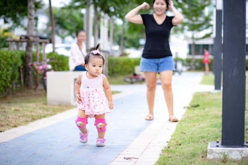 Little baby girl in pink dress and knee pads walking in pavement in park