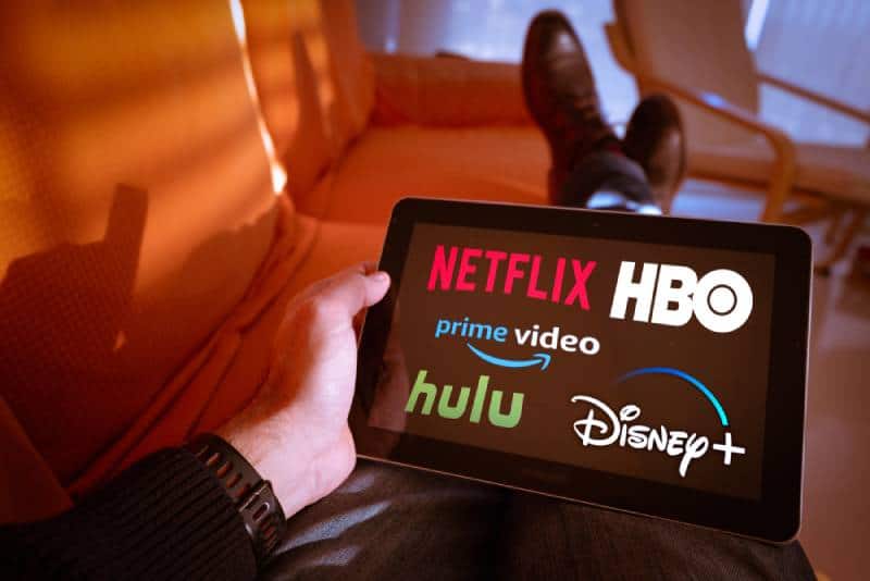 Man holds a tablet with Netflix hulu, amazon video, HBO and Disney+ logos on screen while sitting on a couch