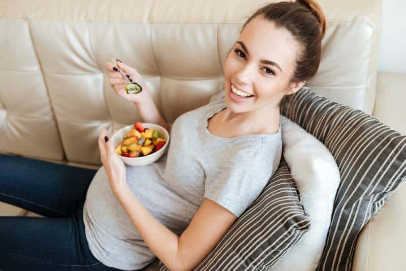 Happy pregnant young woman sitting and eating fruit salad on sofa at home