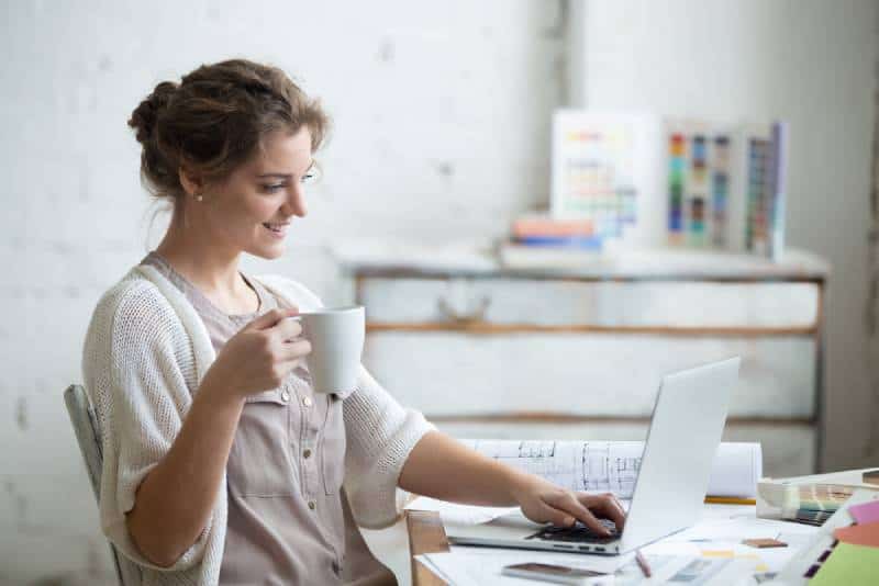 Woman sitting at home office desk with cup of coffee, working on laptop in loft interior