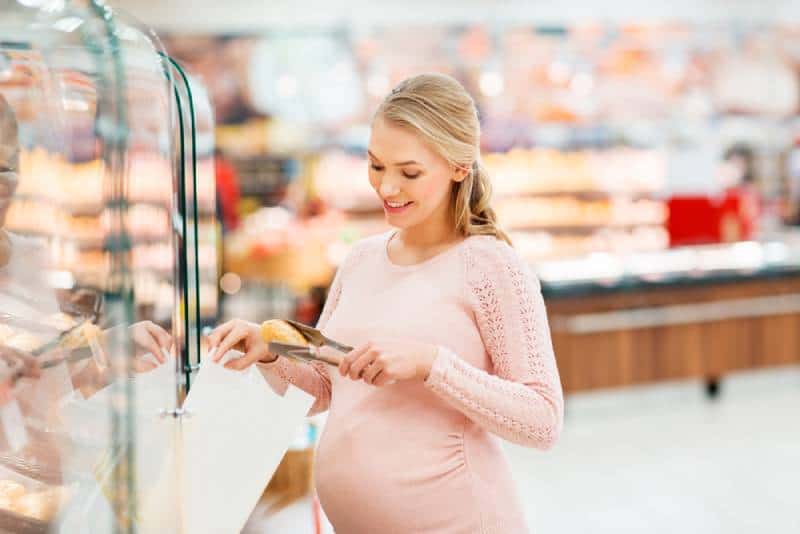 happy pregnant woman with paper bag and tongs buying buns at grocery store or supermarket