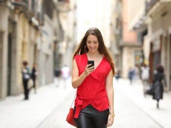 Front view of a fashion happy woman in red shirt and black pants walking and using a smart phone on a city street