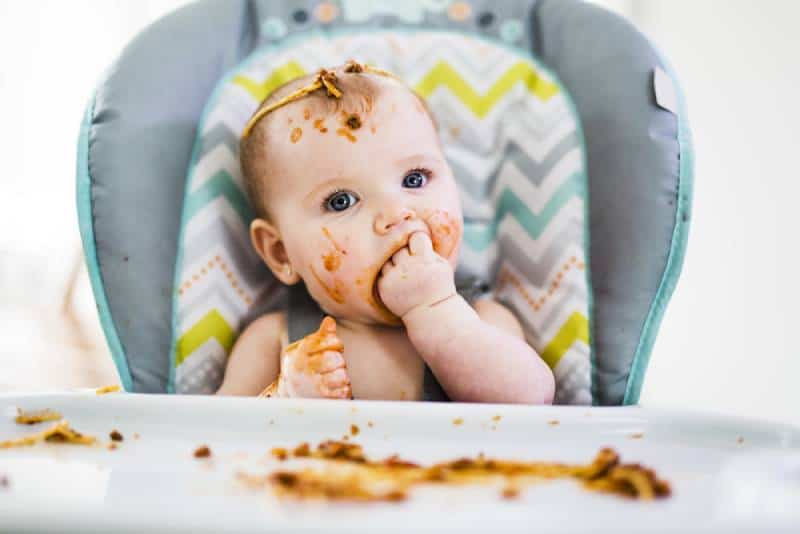 A Little baby eating her dinner and making a mess in a high chair
