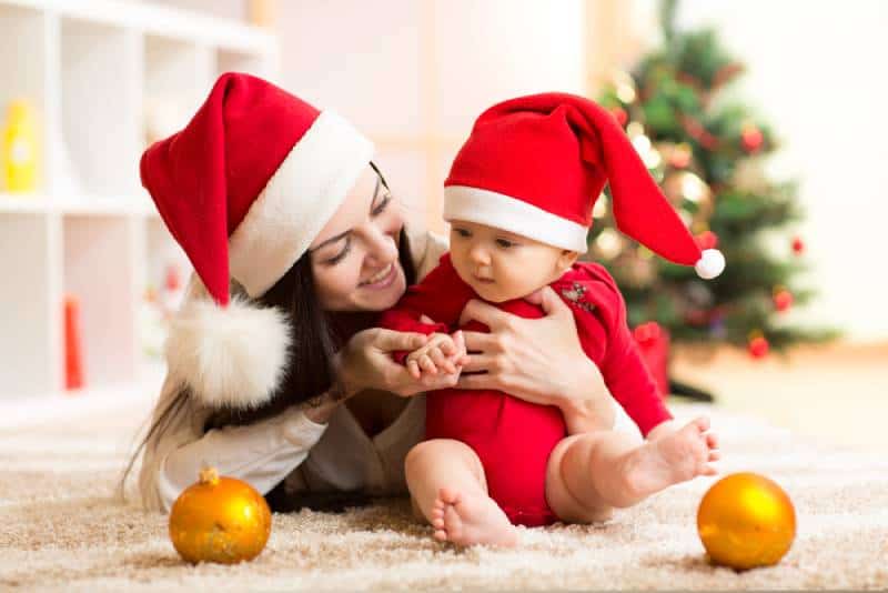Mother and baby in santa red dress smile on a background of Christmas trees in the interior of the house