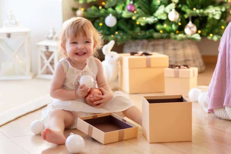 two-year-old girl sits next to an open gift box on the background of the New Year tree and enjoys Christmas toys