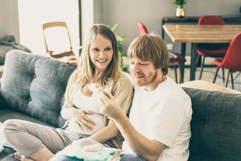 Happy pregnant couple checking baby clothes in living room and smiling