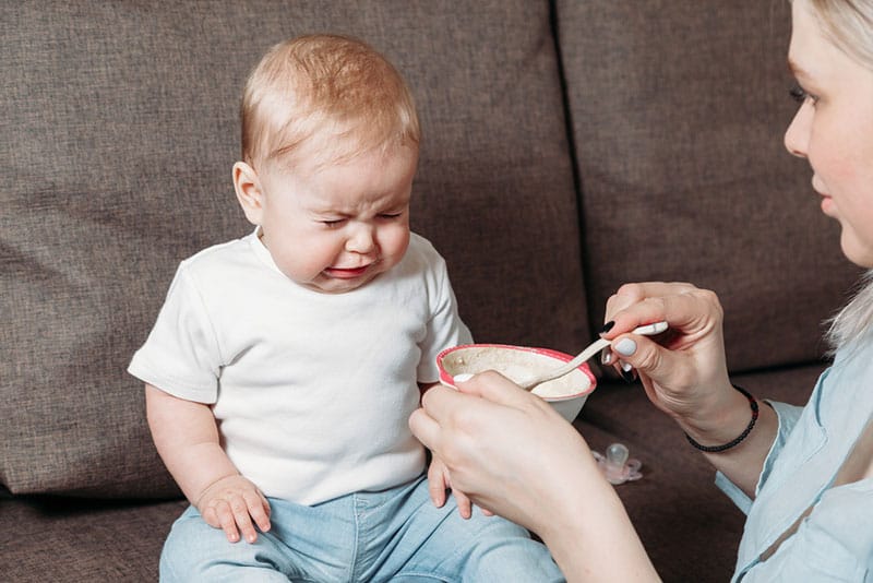 mother trying to feed crying baby on the couch