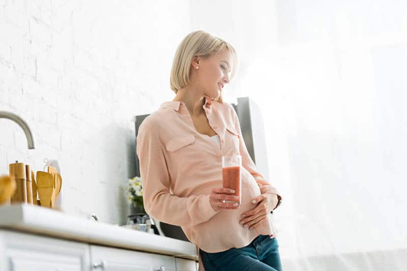 beautiful pregnant woman standing in the kitchen with glass of protein shake
