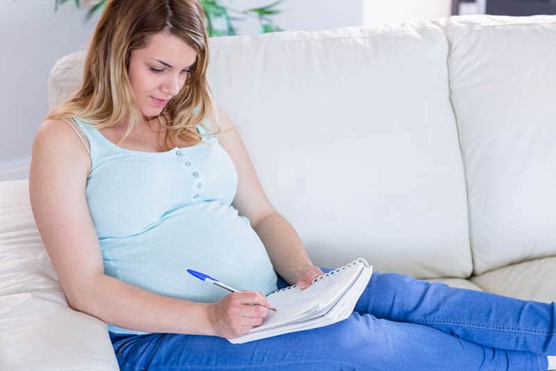 young pregnant woman sitting on the couch and writing a list in notebook