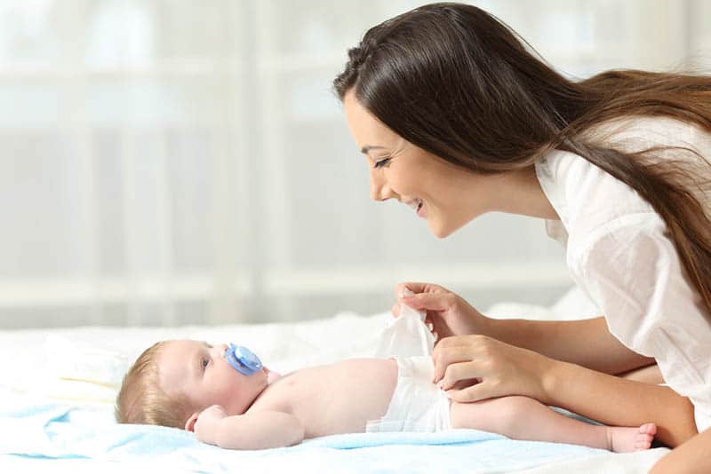 young woman changing diaper to her adorable baby with pacifier on the bed