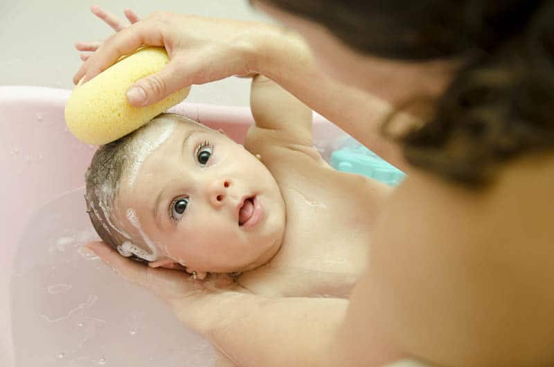 Mutter wäscht Babyhaare mit gelbem Schwamm und einem Shampoo