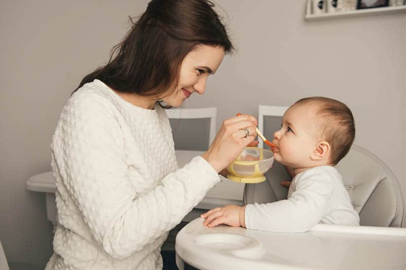 smiling mother feeding baby in high chair with honey cereals