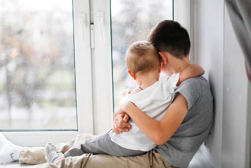 older brother holding in hug his little brother by the window
