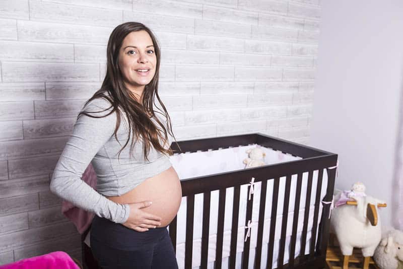 beautiful pregnant woman posing with baby crib in bedroom