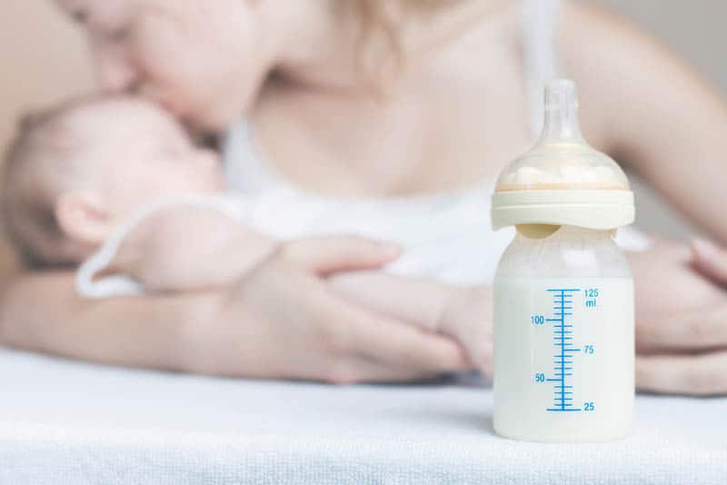 bottle of breast milk and mother kissing a baby in background