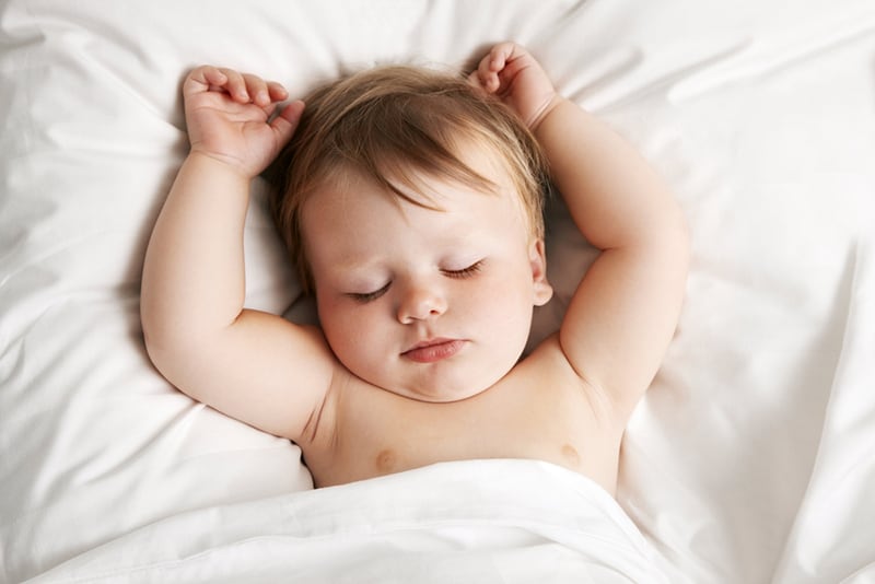 cute baby sleeping on bed in white sheets with hands up
