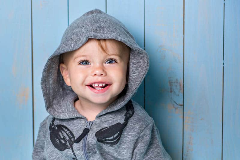 smiling little boy wearing hoodie and standing in front of blue wooden wall