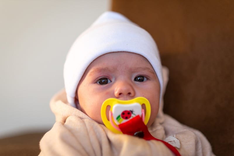 staring baby wearing white winter hat and colorful pacifier